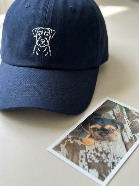 A pet portrait embroidered in white thread on the front of a navy cap, with the owner's own photo of the puppy printed and lying beside it