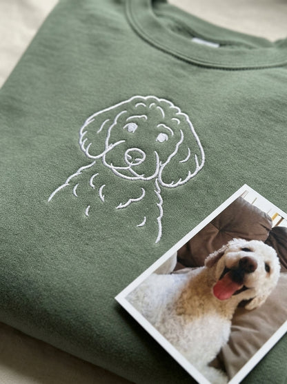 A pet portrait embroidered in white thread on a sage green sweatshirt, with the owner's own photo of the dog printed and lying beside it