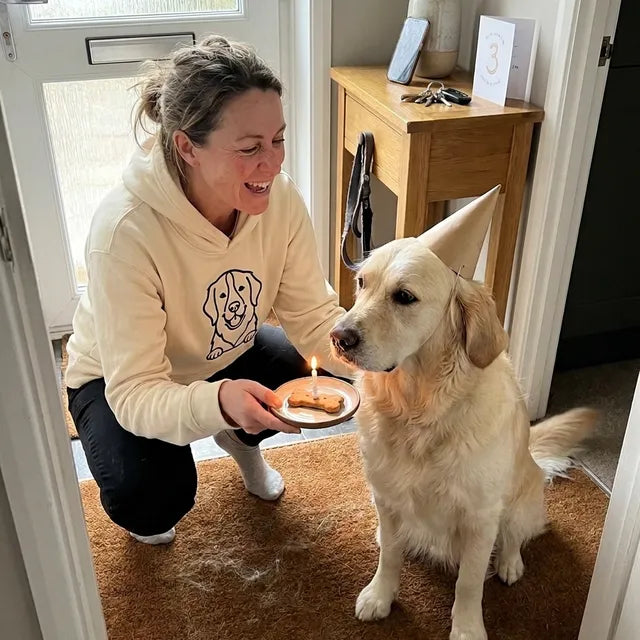A woman holding out a biscuit with a candle to her golden retriever in a party hat